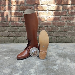 May include: A pair of brown leather riding boots with laces, a round logo, and a detached sole. The boots are tall, reaching the mid-calf, and are set against a brick wall background.