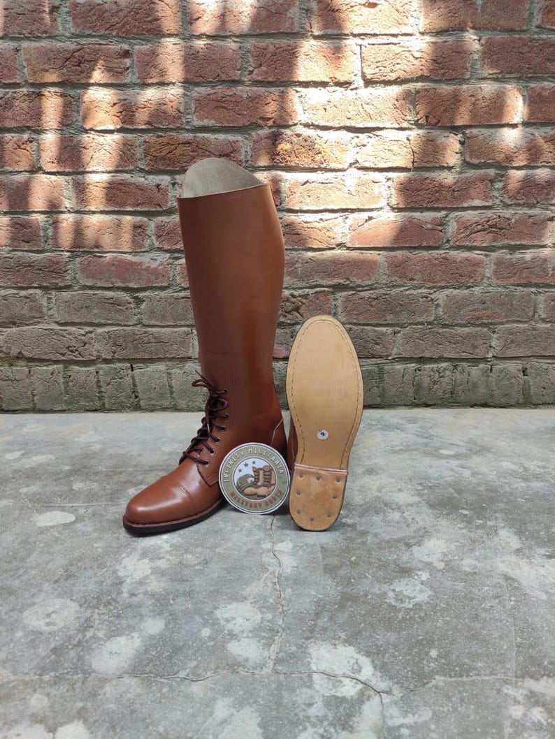 May include: A pair of brown leather riding boots with laces, a round logo, and a detached sole. The boots are tall, reaching the mid-calf, and are set against a brick wall background.