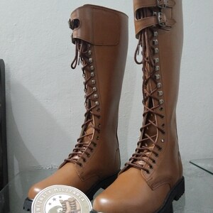 May include: A pair of brown leather knee-high boots with laces and buckles. The boots have a rounded toe and a thick black sole. The boots are displayed on a glass surface. The boots have a military style.