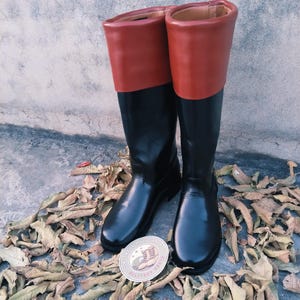 Puede incluir: Botas de cuero negro con la parte superior roja. Las botas son hasta la rodilla y tienen un acabado brillante. Un emblema redondo con texto y un gráfico es visible cerca de las botas. Las botas están sobre hojas secas.