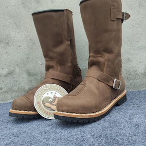 Puede incluir: Botas de cuero marrón con correas de hebilla y suelas negras. Las botas tienen punta redonda y altura media pantorrilla. Se muestra un logotipo circular con "INFINITY MILITARY". Las botas miden aproximadamente 30 cm de alto.