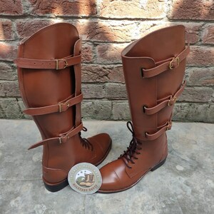 May include: A pair of brown leather riding boots with multiple buckle straps. The boots have a lace-up design and are displayed on a concrete surface against a brick wall. A circular sticker with text and a boot graphic is visible.