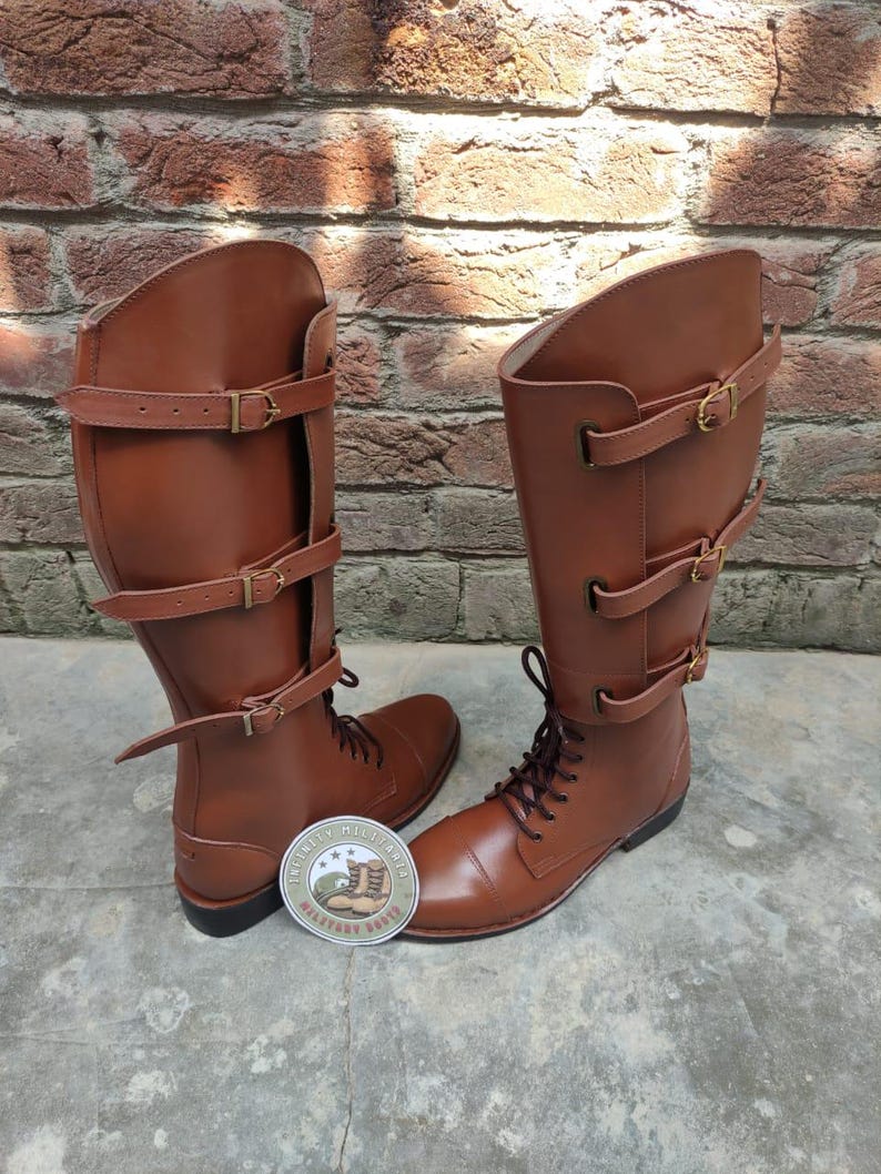 May include: A pair of brown leather riding boots with multiple buckle straps. The boots have a lace-up design and are displayed on a concrete surface against a brick wall. A circular sticker with text and a boot graphic is visible.