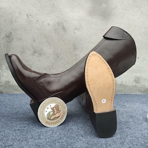 May include: Brown leather riding boots with a rounded toe and a high shaft. The sole is visible, showing a size 7. A circular sticker with text and a boot graphic is next to the boots. The boots are on a gray surface.