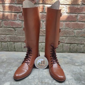 May include: A pair of tall, brown leather boots with laces and buckles. The boots have a classic design and are standing upright. A round sticker with text and a boot graphic is between the boots.