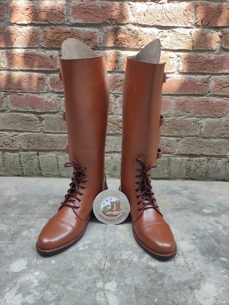 May include: A pair of tall, brown leather boots with laces and buckles. The boots have a classic design and are standing upright. A round sticker with text and a boot graphic is between the boots.