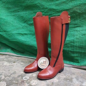 Puede incluir: Un par de botas altas de cuero marrón rojizo. Las botas tienen una cremallera lateral, un panel negro y un tacón pequeño. Una pegatina redonda con texto y un logotipo se coloca entre las botas. El fondo es de tela verde.