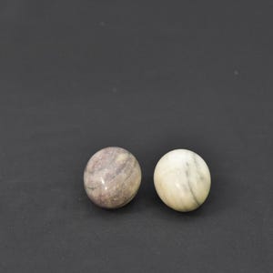 May include: Two marble spheres, one a mottled mix of gray and pink, the other a creamy white with subtle gray veining. The spheres are smooth and polished, set against a dark gray background, showcasing their natural textures and colors.