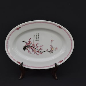May include: White oval serving platter with a pink floral design and a decorative border. The platter features a branch with pink blossoms and Chinese characters. The platter is displayed on a dark brown stand.