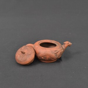 Op de afbeelding: Kleine, terracotta-kleurige theepot met een drakenontwerp. Het deksel is eraf, waardoor de binnenkant zichtbaar is. De tuit is gevormd als een drakenkop. De theepot is waarschijnlijk van klei.