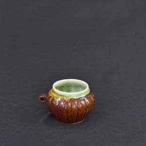 May include: A small, brown ceramic cup with a handle. The cup has a ribbed texture and a green interior. The rim is accented with yellow and green glaze. The cup is likely for decorative purposes.