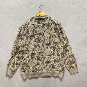 Puede incluir: Sudadera beige de manga larga con un estampado de camuflaje marr&oacute;n. La sudadera tiene cuello y pu&ntilde;os y dobladillo de canal&eacute;. El texto es visible en la parte posterior del cuello. La prenda est&aacute; colgada de una percha de madera.