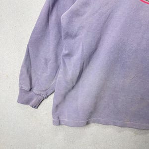 May include: A faded lavender long-sleeved sweatshirt. The cuffs and hem are ribbed. The fabric has a worn appearance. The upper right corner of the sweatshirt has a partial design with pink and green stripes.
