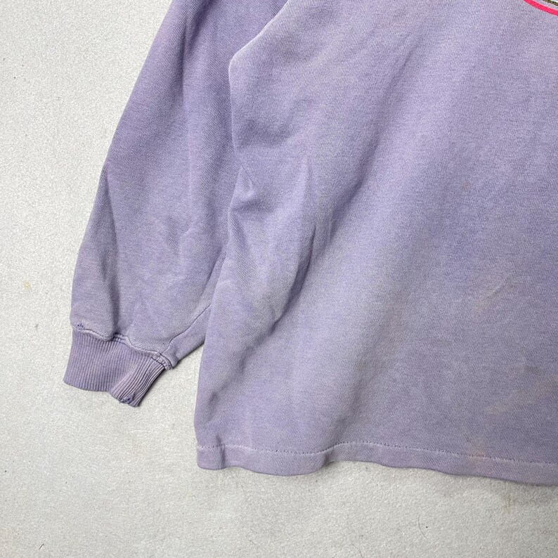 May include: A faded lavender long-sleeved sweatshirt. The cuffs and hem are ribbed. The fabric has a worn appearance. The upper right corner of the sweatshirt has a partial design with pink and green stripes.