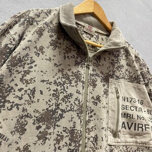 Puede incluir: Chaqueta con cremallera de camuflaje marr&oacute;n y beige con cuello de pana. La chaqueta tiene un bolsillo delantero con el texto "N173-18 SECT/R-8L MRI. No. 22 AVIREX". La chaqueta est&aacute; colgada en una percha de madera.