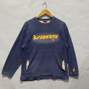 以下が含まれることがあります： 胸に黄色の「Vans」の文字が入った、ネイビーブルーの長袖スウェットシャツ。スウェットシャツには両サイドにポケットがあり、袖には小さなロゴが付いています。衣類は木製のハンガーに吊るされています。