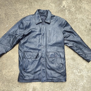 Könnte beinhalten: Eine marineblaue Lederjacke mit Kragen, Knopfverschluss und zwei Vordertaschen. Die Jacke hat ein Steppmuster und ein klassisches Design. Die Jacke liegt flach auf einer Betonoberfläche.