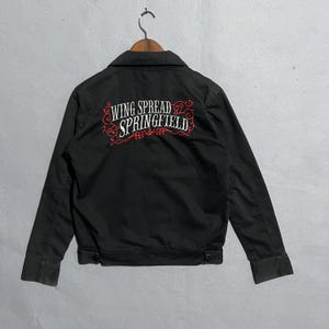 Puede incluir: Chaqueta negra con el texto bordado "WING SPREAD SPRINGFIELD" en blanco y rojo. La chaqueta tiene cuello, mangas largas y dos cierres de botón en la cintura. La chaqueta está colgada en una percha de madera.