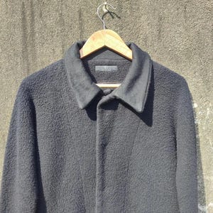 May include: A black wool coat with a textured finish hangs on a wooden hanger. The coat features a classic collar and a button-down front. The coat is displayed against a neutral-coloured wall.