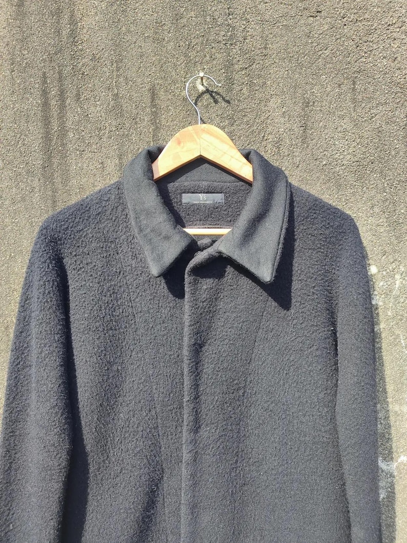 May include: A black wool coat with a textured finish hangs on a wooden hanger. The coat features a classic collar and a button-down front. The coat is displayed against a neutral-coloured wall.