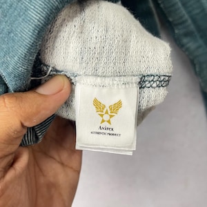 Peut inclure: Gros plan sur l'int&eacute;rieur d'un v&ecirc;tement bleu sarcelle, r&eacute;v&eacute;lant une doublure blanche textur&eacute;e. Une &eacute;tiquette rectangulaire avec le logo Avirex, une &eacute;toile ail&eacute;e et le texte "AUTHENTIC PRODUCT" est visible.