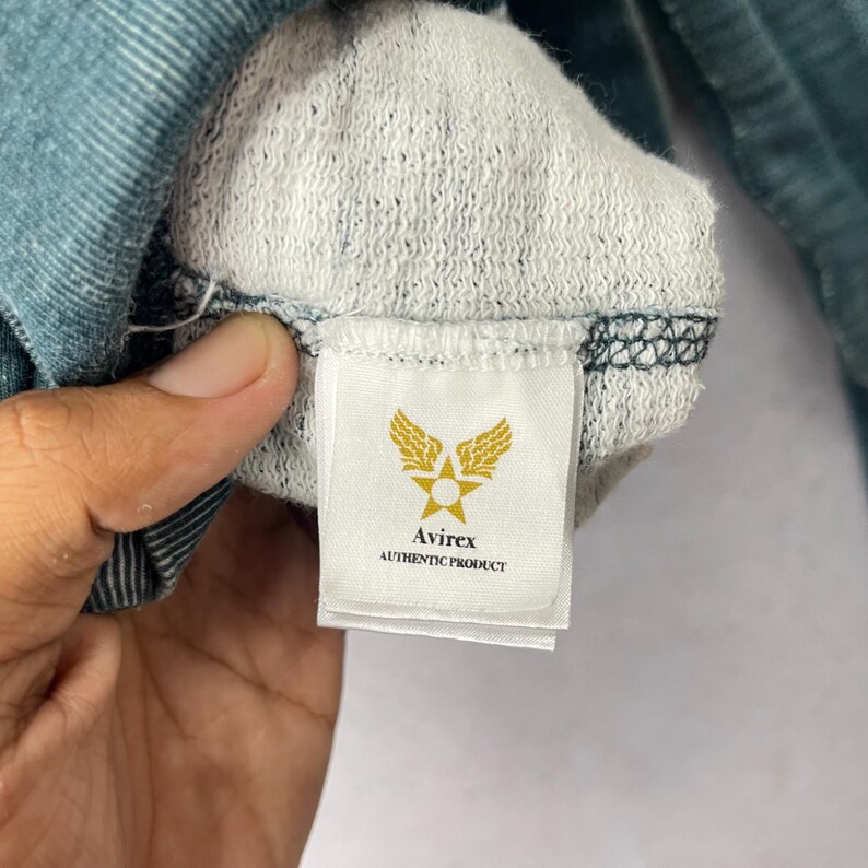 Peut inclure: Gros plan sur l'int&eacute;rieur d'un v&ecirc;tement bleu sarcelle, r&eacute;v&eacute;lant une doublure blanche textur&eacute;e. Une &eacute;tiquette rectangulaire avec le logo Avirex, une &eacute;toile ail&eacute;e et le texte "AUTHENTIC PRODUCT" est visible.