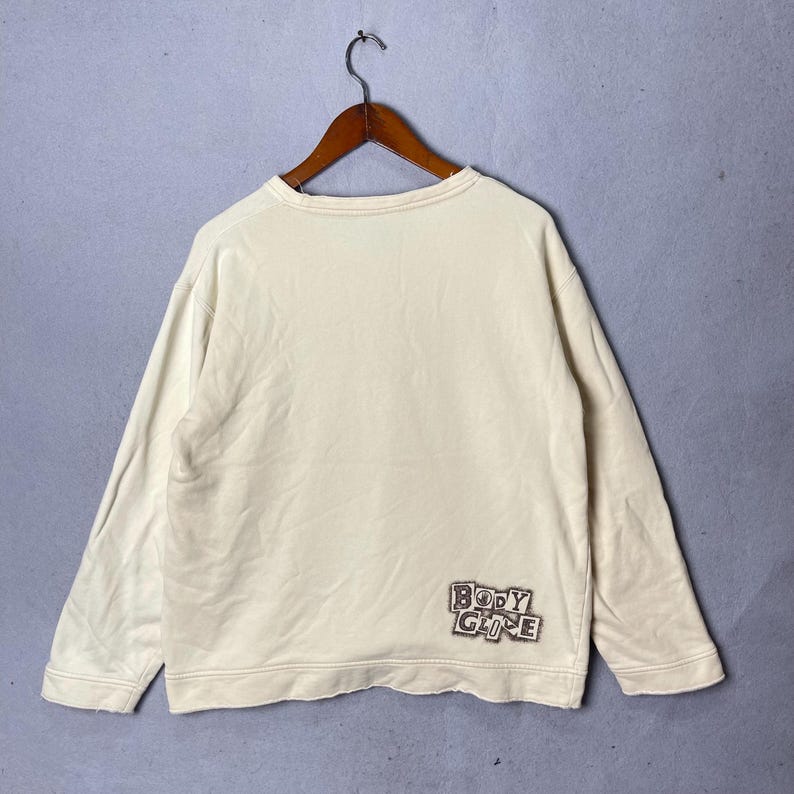 Puede incluir: Una sudadera de manga larga de color crema con cuello redondo y pu&ntilde;os acanalados. La prenda presenta un parche gr&aacute;fico rectangular en la parte inferior derecha con el texto "BODY GLIDE" en una fuente negra estilizada y desgastada.