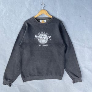 Op de afbeelding: Een grijze Hard Rock Cafe Hollywood sweatshirt met lange mouwen. De voorkant heeft het Hard Rock Cafe logo in wit, met de woorden "SAVE THE PLANET" erboven en "HOLLYWOOD" eronder. De sweatshirt hangt aan een houten hanger.