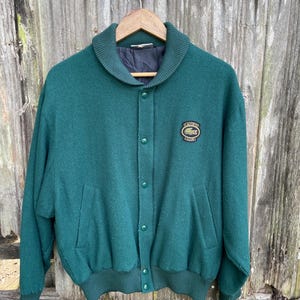 May include: A dark green wool jacket with a button-up front, a wide collar, and a ribbed hem and cuffs. The jacket features a small embroidered patch on the chest. The jacket is hanging on a wooden hanger against a weathered wooden background.