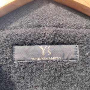 May include: Close-up of a dark grey garment with a textured, fuzzy surface. A navy blue label is sewn onto the garment, displaying the text "Y's YOHJI YAMAMOTO" in a gold colour.