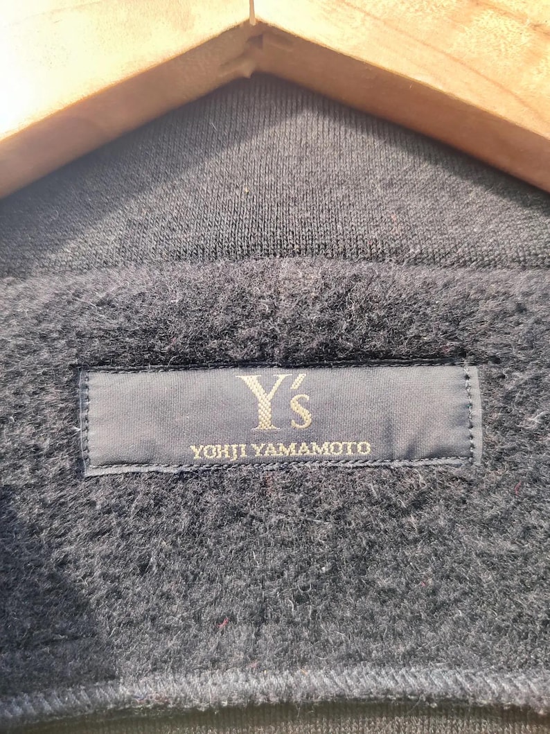 May include: Close-up of a dark grey garment with a textured, fuzzy surface. A navy blue label is sewn onto the garment, displaying the text "Y's YOHJI YAMAMOTO" in a gold colour.