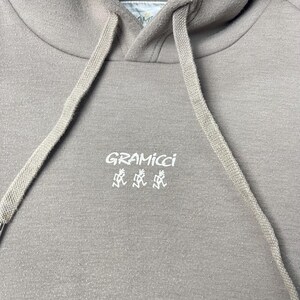 May include: A close-up of a taupe hoodie with a drawstring and a hood. The white text "GRAMICCI" and a stylized graphic are printed on the front. The hoodie is made of a soft, textured fabric.