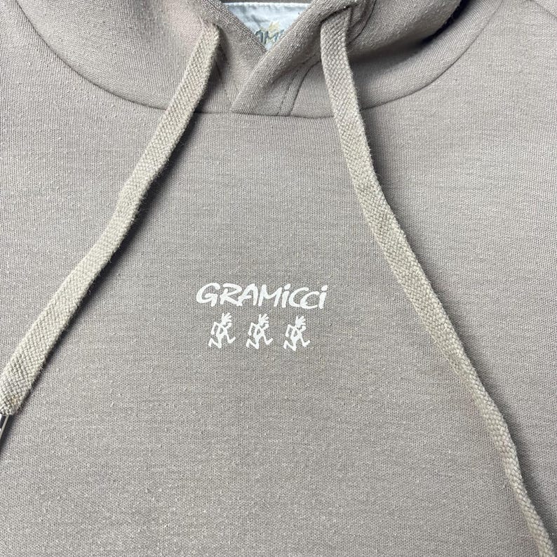 May include: A close-up of a taupe hoodie with a drawstring and a hood. The white text "GRAMICCI" and a stylized graphic are printed on the front. The hoodie is made of a soft, textured fabric.