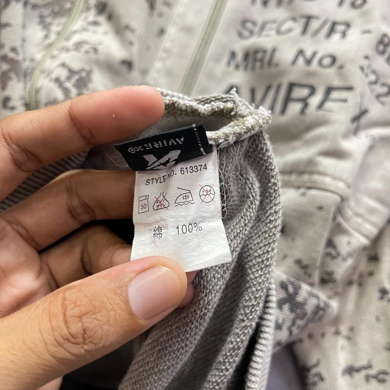 Puede incluir: Primer plano de una etiqueta de ropa de una sudadera con cremallera Avirex. La etiqueta muestra el logotipo de la marca, el n&uacute;mero de estilo 613374 e instrucciones de cuidado. La sudadera tiene un patr&oacute;n de camuflaje gris y blanco.