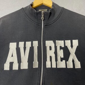 Puede incluir: Una sudadera negra con cremallera con el texto bordado "AVI REX" en gris claro, con una fuente de estilo universitario. La sudadera tiene un cuello alto y una cremallera plateada en la parte delantera.