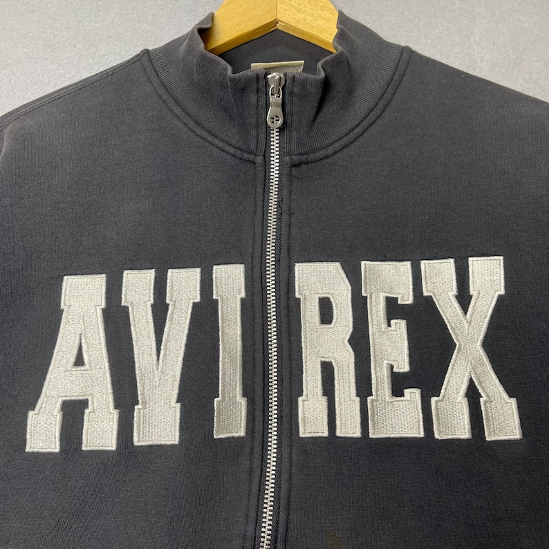 Puede incluir: Una sudadera negra con cremallera con el texto bordado "AVI REX" en gris claro, con una fuente de estilo universitario. La sudadera tiene un cuello alto y una cremallera plateada en la parte delantera.