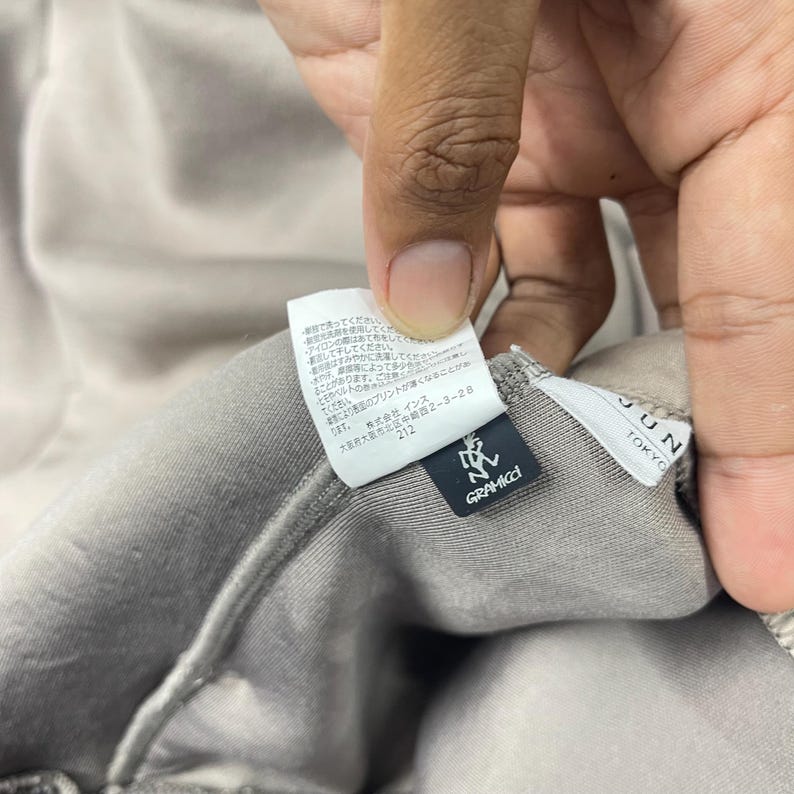 May include: Close-up of a gray fabric with a tag attached. The tag has Japanese text and the brand name "GRAMICCI" in white. The tag also has the text "JUN TOKYO" in white. The fabric is a solid gray color.