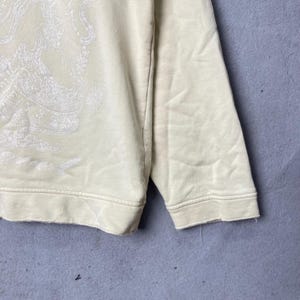 Puede incluir: Una sudadera de manga larga color crema con un gran estampado gr&aacute;fico blanco e intrincado en la parte delantera. La prenda tiene cuello redondo, mangas largas con pu&ntilde;os acanalados y un dobladillo sin rematar en el borde inferior.