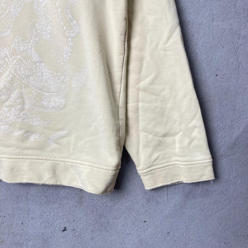 Puede incluir: Una sudadera de manga larga color crema con un gran estampado gr&aacute;fico blanco e intrincado en la parte delantera. La prenda tiene cuello redondo, mangas largas con pu&ntilde;os acanalados y un dobladillo sin rematar en el borde inferior.