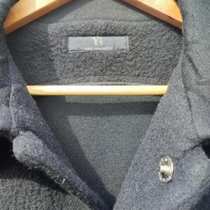 May include: Close-up of a dark grey jacket with a button fastening. The jacket features a textured fabric and a label reading "Y's YOHJI YAMAMOTO". The jacket is suspended on a wooden hanger.