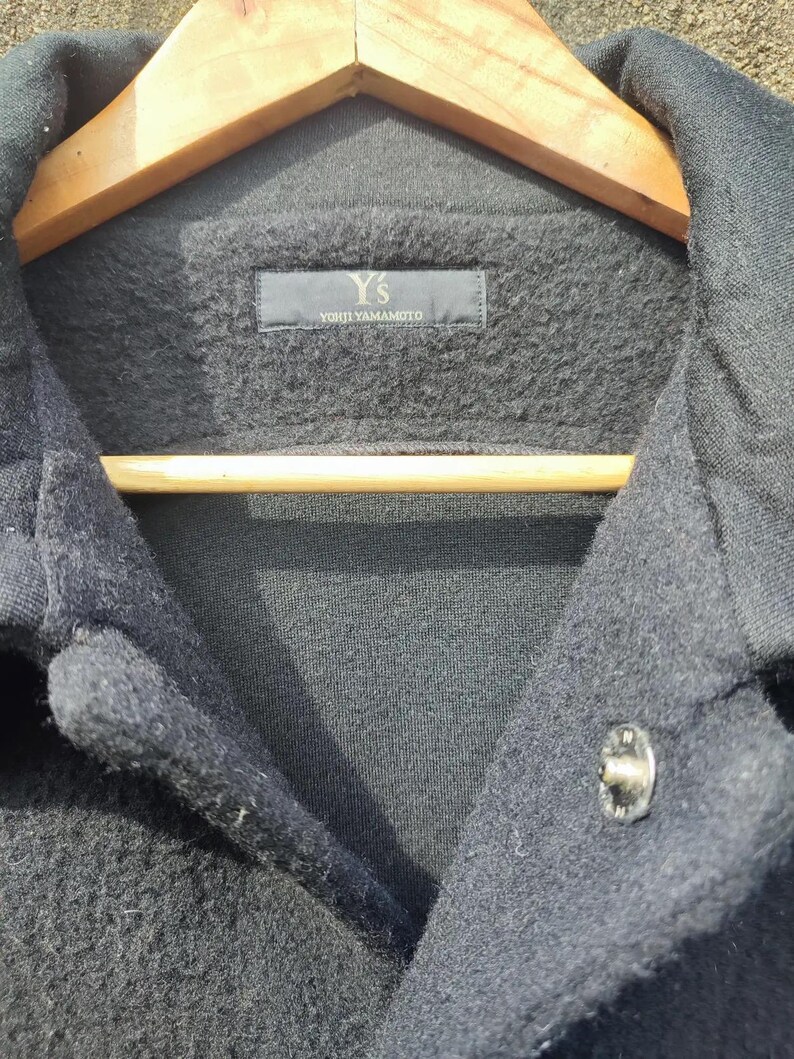 May include: Close-up of a dark grey jacket with a button fastening. The jacket features a textured fabric and a label reading "Y's YOHJI YAMAMOTO". The jacket is suspended on a wooden hanger.