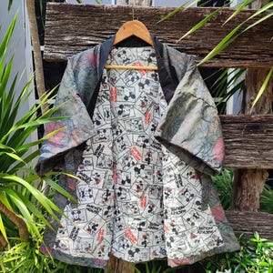 Może przedstawiać: Wzorzysta kurtka w stylu kimono z czarnym kołnierzem i podszewką z nadrukiem Myszki Miki. Zewnętrzny materiał ma kwiatowy wzór w odcieniach szarości, różu i zieleni. Kurtka wisi na drewnianym wieszaku na tle rustykalnego drewnianego tła.