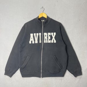 Puede incluir: Una sudadera negra con cremallera y el texto blanco "AVI REX" en el pecho. La sudadera tiene cuello alto, dos bolsillos delanteros y una cremallera plateada. La prenda est&aacute; colgada de una percha de madera sobre un fondo gris.