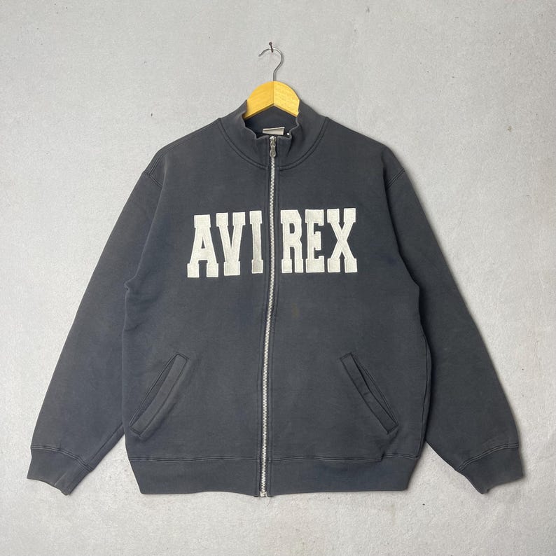 Puede incluir: Una sudadera negra con cremallera y el texto blanco "AVI REX" en el pecho. La sudadera tiene cuello alto, dos bolsillos delanteros y una cremallera plateada. La prenda est&aacute; colgada de una percha de madera sobre un fondo gris.