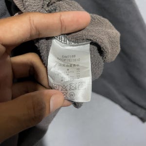 Puede incluir: Primer plano de una etiqueta de prenda gris con texto y s&iacute;mbolos. La etiqueta incluye los n&uacute;meros "DM7168" y "MCF(5) 1810". La etiqueta tambi&eacute;n tiene texto japon&eacute;s e instrucciones de lavado.