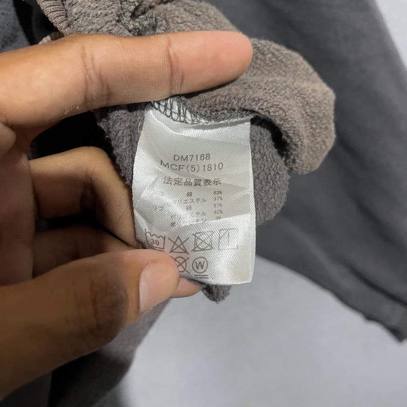 Puede incluir: Primer plano de una etiqueta de prenda gris con texto y s&iacute;mbolos. La etiqueta incluye los n&uacute;meros "DM7168" y "MCF(5) 1810". La etiqueta tambi&eacute;n tiene texto japon&eacute;s e instrucciones de lavado.