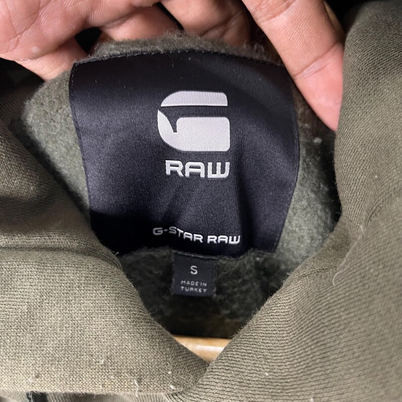 Puede incluir: Primer plano del cuello de una sudadera con capucha verde con una etiqueta negra. La etiqueta presenta el texto blanco "RAW" encima de "G-STAR RAW". Debajo, una peque&ntilde;a etiqueta dice "S" y "MADE IN TURKEY".