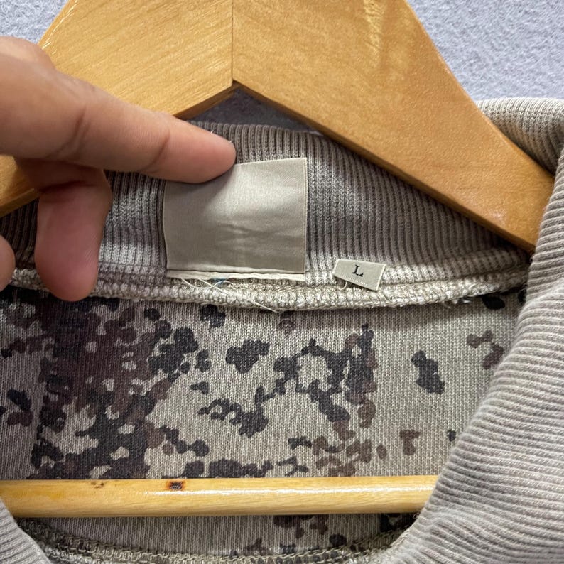 Puede incluir: Primer plano de un cuello de sudadera gris con forro de estampado de camuflaje. El cuello est&aacute; en una percha de madera. Es visible una peque&ntilde;a etiqueta con la letra "L".