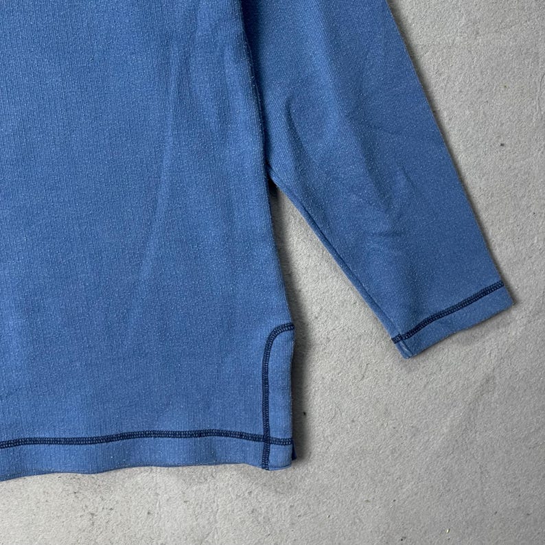 Puede incluir: Una camisa azul claro de manga larga con costuras azul oscuro a lo largo de las costuras. La camisa tiene un dise&ntilde;o sencillo y est&aacute; hecha de una tela suave y texturizada. Las mangas son de tres cuartos.
