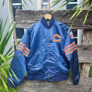Puede incluir: Chaqueta bomber de satén azul marino con rayas naranjas y blancas en las mangas y el logotipo de los Chicago Bears en el pecho. La chaqueta tiene una parte delantera con botones y un cuello y puños azul marino.